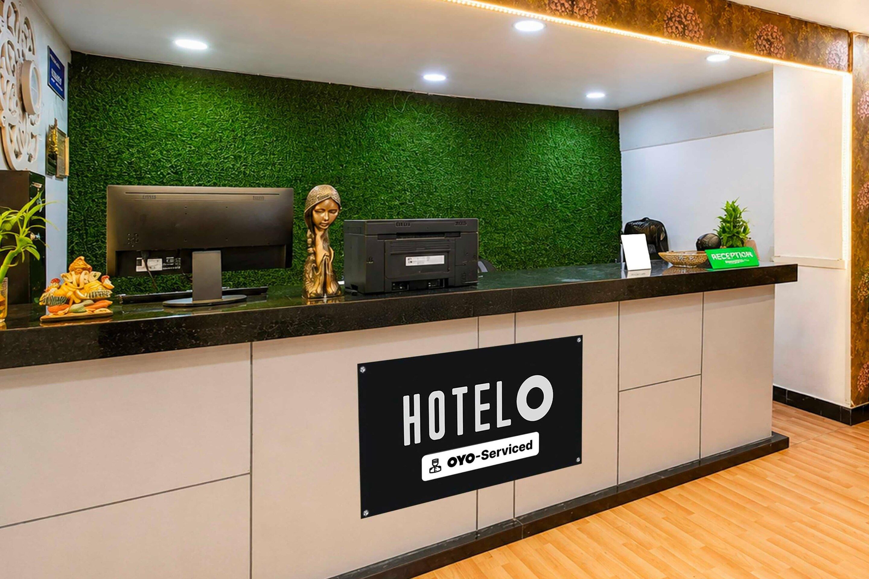 Balcão de recepção de hotel com parede verde, placa de recepção, computador e impressora.