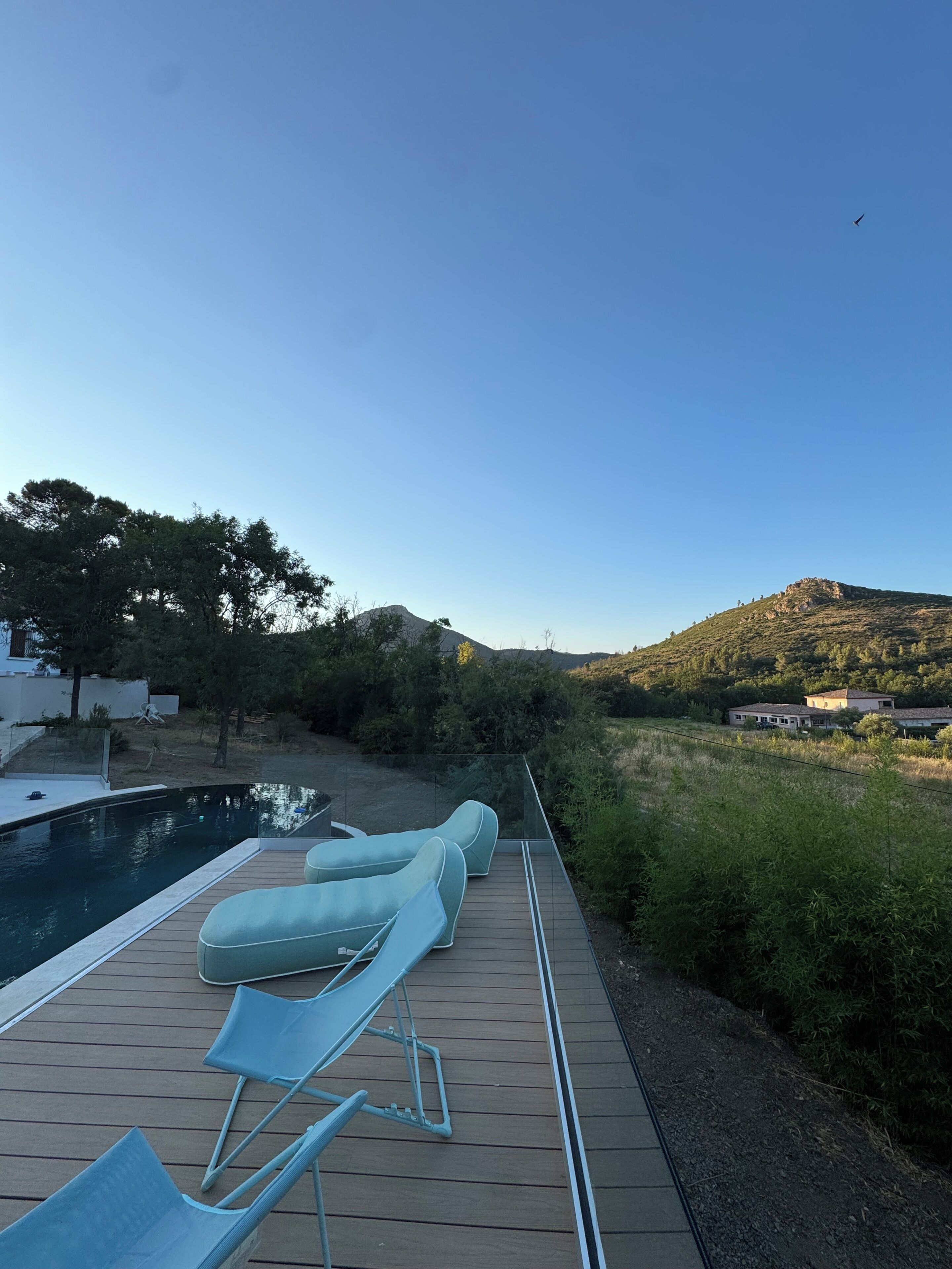 A deck with lounge chairs overlooking a body of water, a pool, and a hilly landscape.