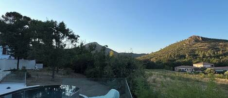 A deck with lounge chairs overlooking a body of water, a pool, and a hilly landscape.