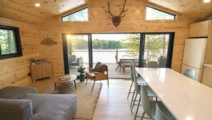 A wooden cabin interior with a dining area, sofa, and a large window overlooking a body of water. - Scandinavian Cabin on Sandy Lakefront [NEW RENOVATION] (Lake Ann)