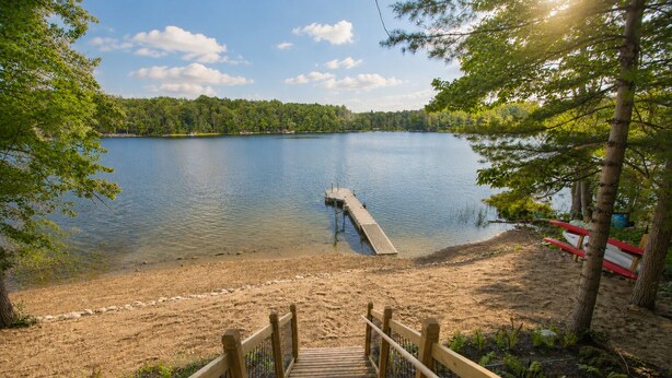 A wooden deck leading to a sandy beach with a pier extending into a calm lake, surrounded by trees. - Scandinavian Cabin on Sandy Lakefront [NEW RENOVATION] (Lake Ann)