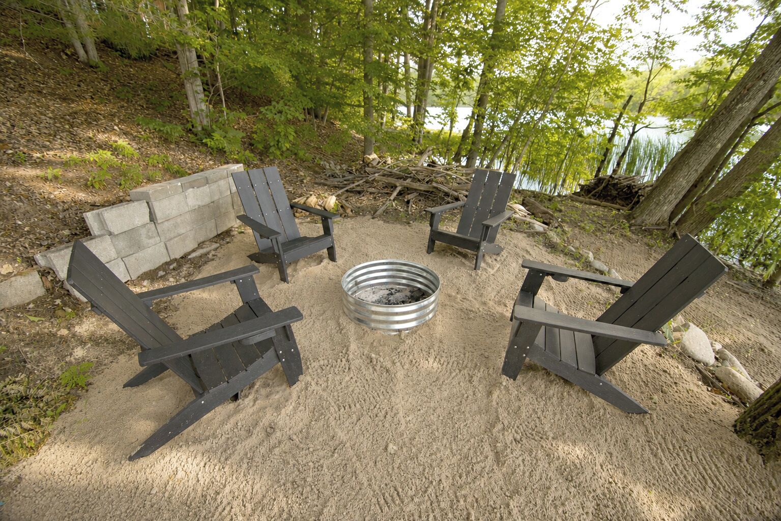 Disposición circular de asientos con cuatro sillas Adirondack alrededor de un brasero de metal en un entorno natural.