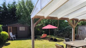 Een overdekt terras met een tafel en stoelen, een rode parasol en een houten schuurtje op de achtergrond.