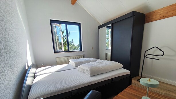 Een compacte slaapkamer met een bed, bureau en stoel. Er is een raam met uitzicht op bomen en een gebouw.