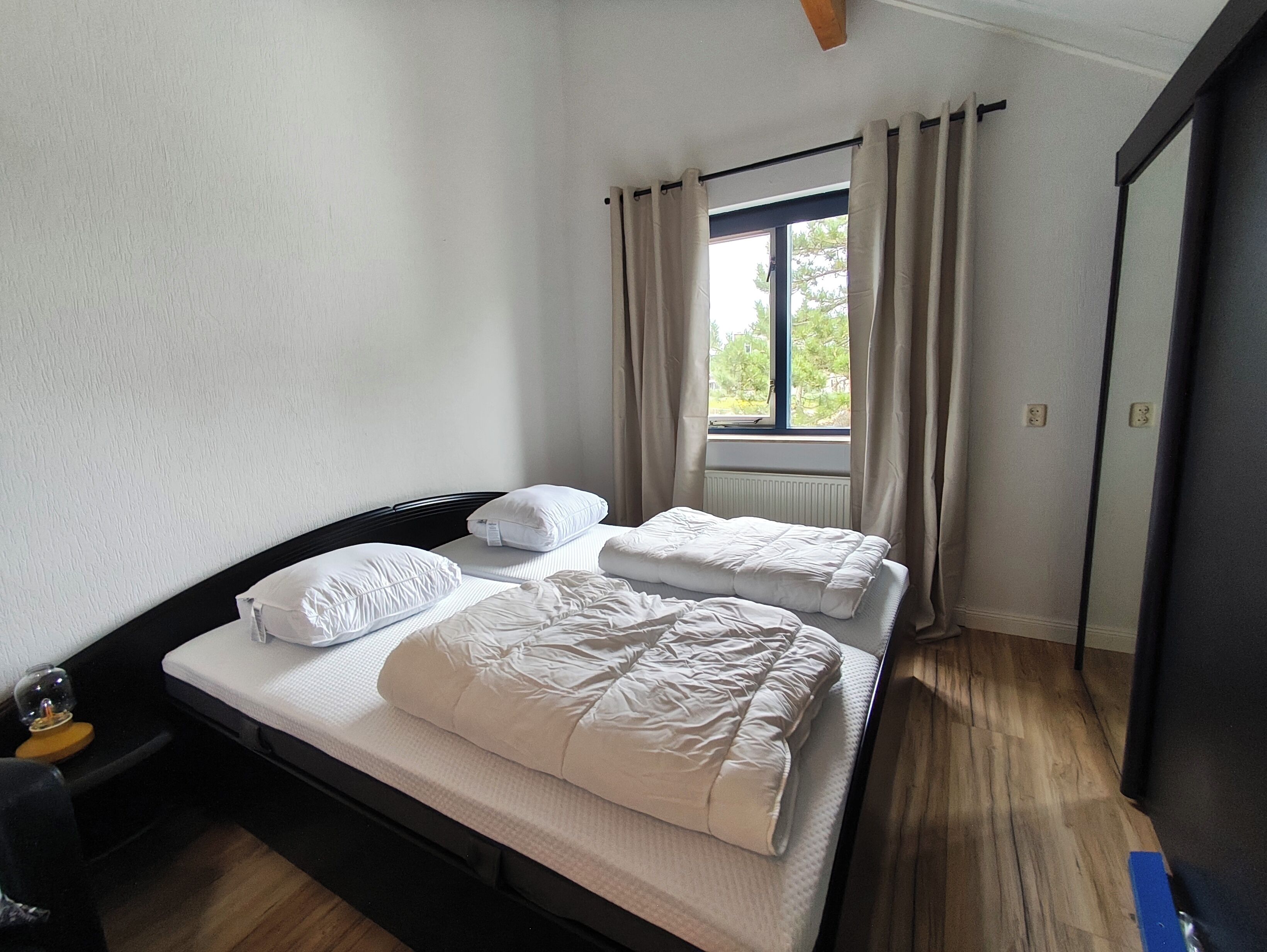 Een slaapkamer met een bed, twee kussens, een raam met gordijnen en een radiator.