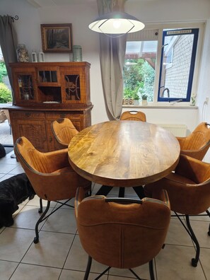 A dining area with a round wooden table surrounded by six brown chairs, a wooden cabinet with glass doors, a painting on the wall, and a window with a view of greenery. - Cozy vacation home near the water—a relaxing getaway for people and their dog(s)! (Julianadorp)