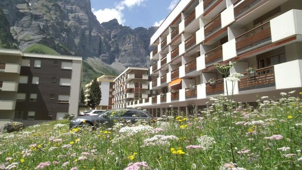 Ett modernt hotell med flera balkonger, omgivet av ett bergigt landskap och en äng full av vilda blommor.