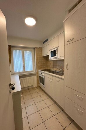 Eine kleine Küche mit weißen Schränken, einer Mikrowelle, einem Spülbecken und einem Fenster mit Jalousien.