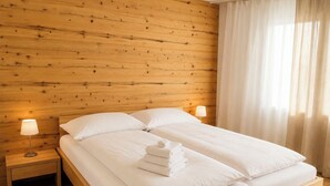 A hotel room with a wooden headboard, a double bed with white linens, two bedside tables with lamps, and a window with sheer curtains. - Majesta Leukerbad # 6-bed apartment, Dusche, WC by Interhome (Leukerbad)