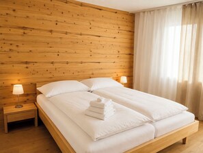 A hotel room with a wooden headboard, a double bed with white linens, two bedside tables with lamps, and a window with sheer curtains. - Majesta Leukerbad # 6-bed apartment, Dusche, WC by Interhome (Leukerbad)