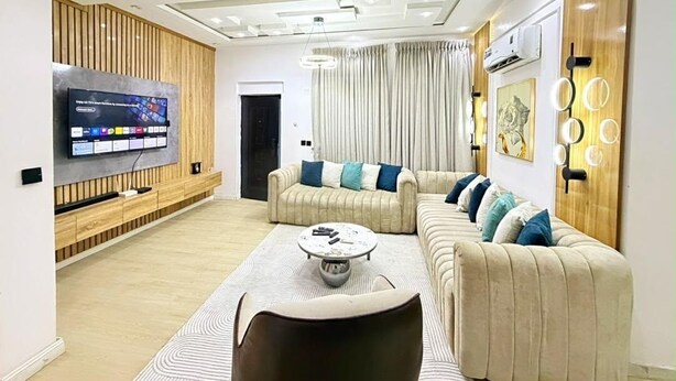 A modern living room with a sectional sofa, a coffee table, and a flat-screen TV mounted on the wall. - Beautifully Styled 2 Bedroom Apartment in Salem Ikate. QQ (Ikate)