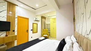 A modern hotel room with a large bed, a flat-screen TV, a wooden wardrobe, and a bathroom with a yellow chair. - Beautifully Styled 2 Bedroom Apartment in Salem Ikate. QQ (Ikate)
