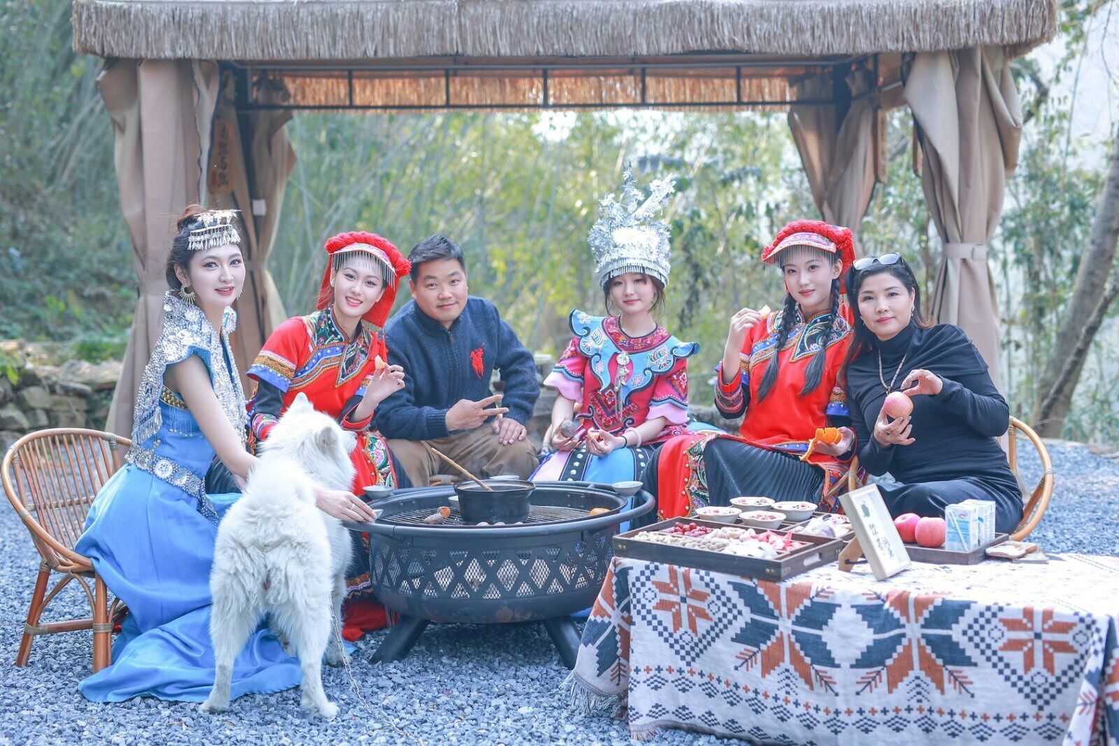 Un grupo de personas vestidas con atuendos tradicionales sentadas alrededor de una estructura de techo de paja, con una parrilla, un perro y una mesa con comida.