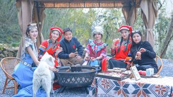 Un grupo de personas vestidas con atuendos tradicionales sentadas alrededor de una estructura de techo de paja, con una parrilla, un perro y una mesa con comida.