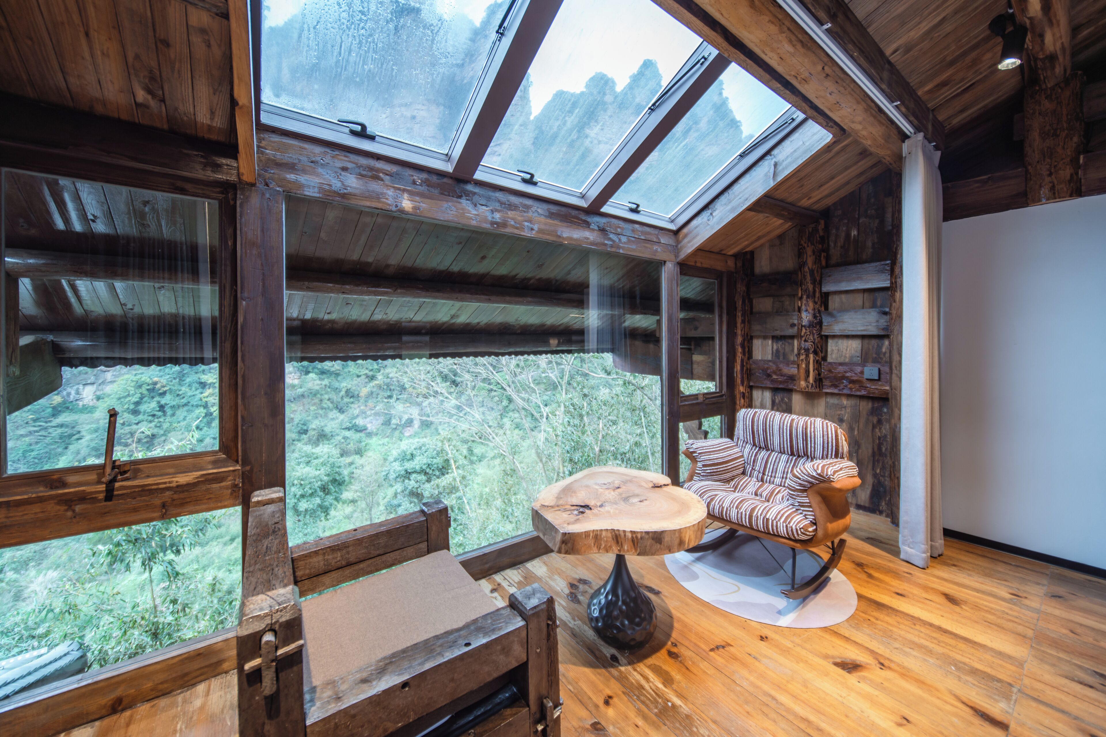 Interior de una cabaña de madera con un tragaluz grande, una mesa redonda de madera, una silla a rayas y un florero.