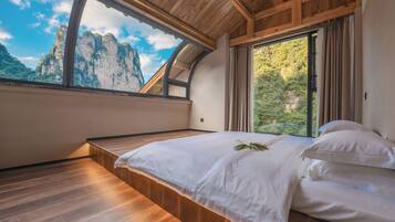 A bedroom with a wooden ceiling, a large window with a view of mountains, a bed with white linens, and a wooden floor.