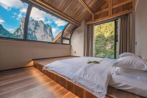 Un dormitorio con techo de madera, un ventanal con vistas a montañas, una cama con sábanas blancas y piso de madera.