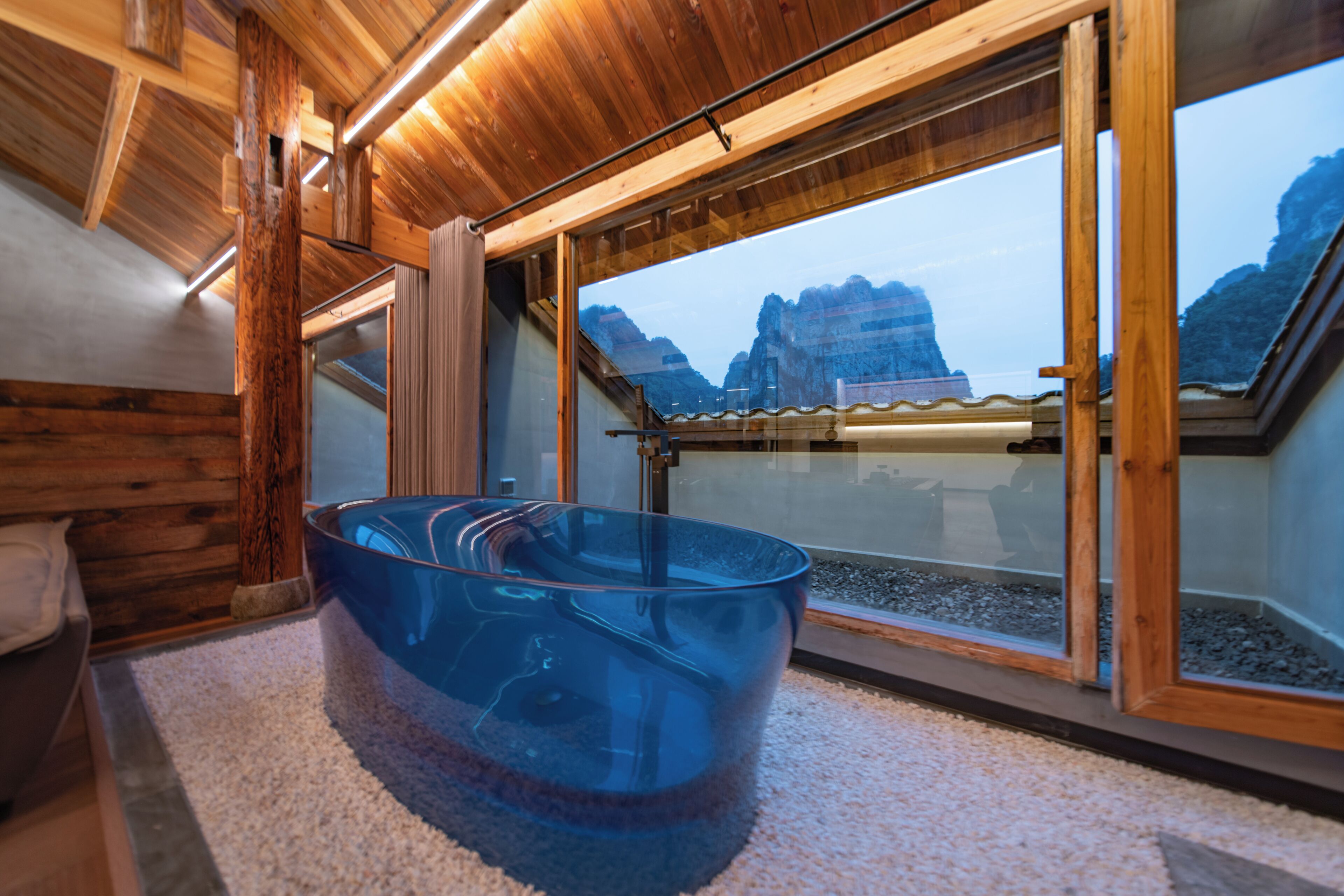 Un interior moderno con una bañera de vidrio, techo de madera y grandes ventanas con vistas a la montaña.