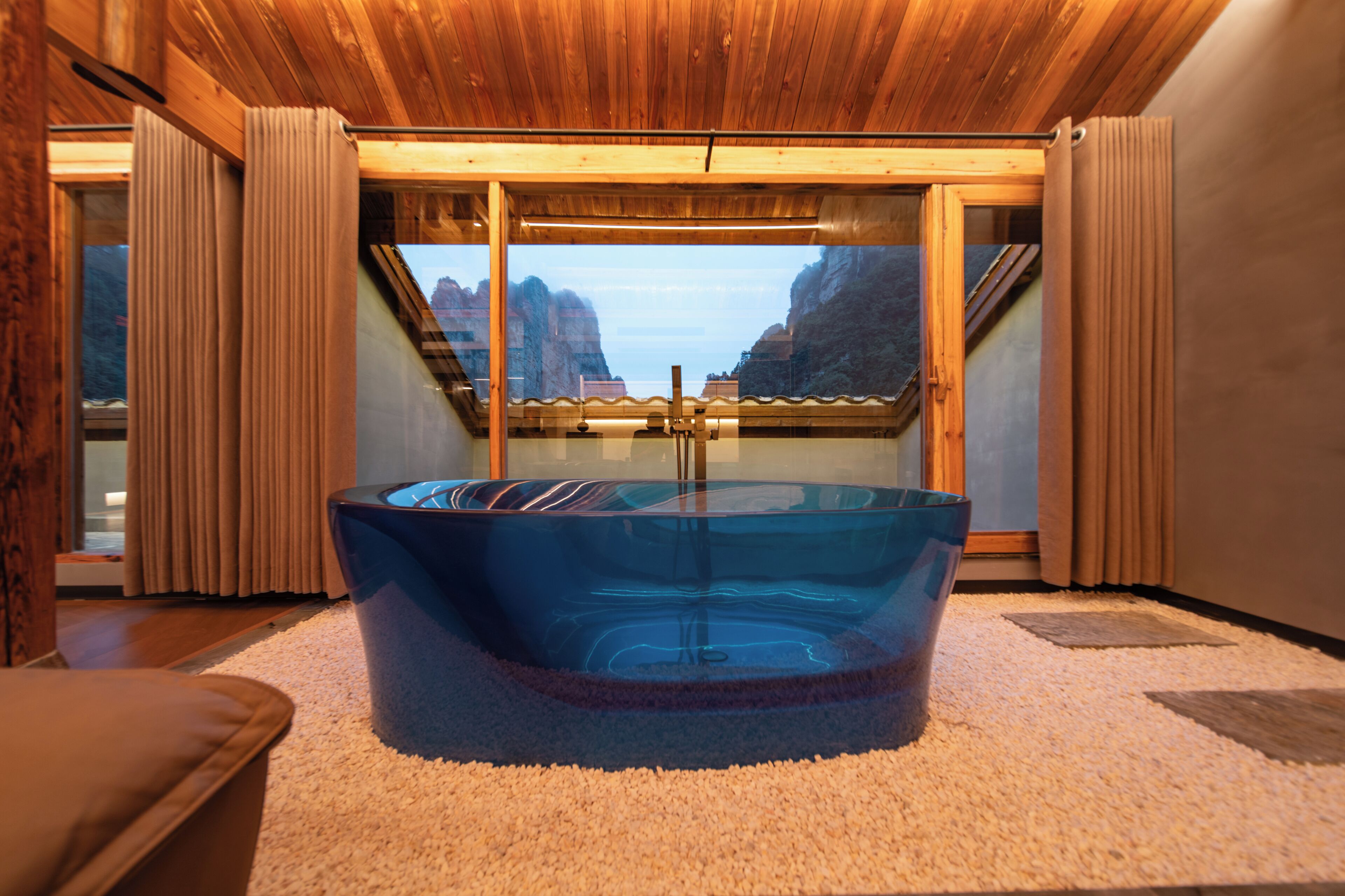 Habitación con una bañera de vidrio grande, techo de madera y vistas a las montañas a través de un ventanal.