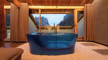 Ein Zimmer mit einer großen Eckbadewanne aus Glas, Holzboden und Blick auf Berge durch eine Glastür.