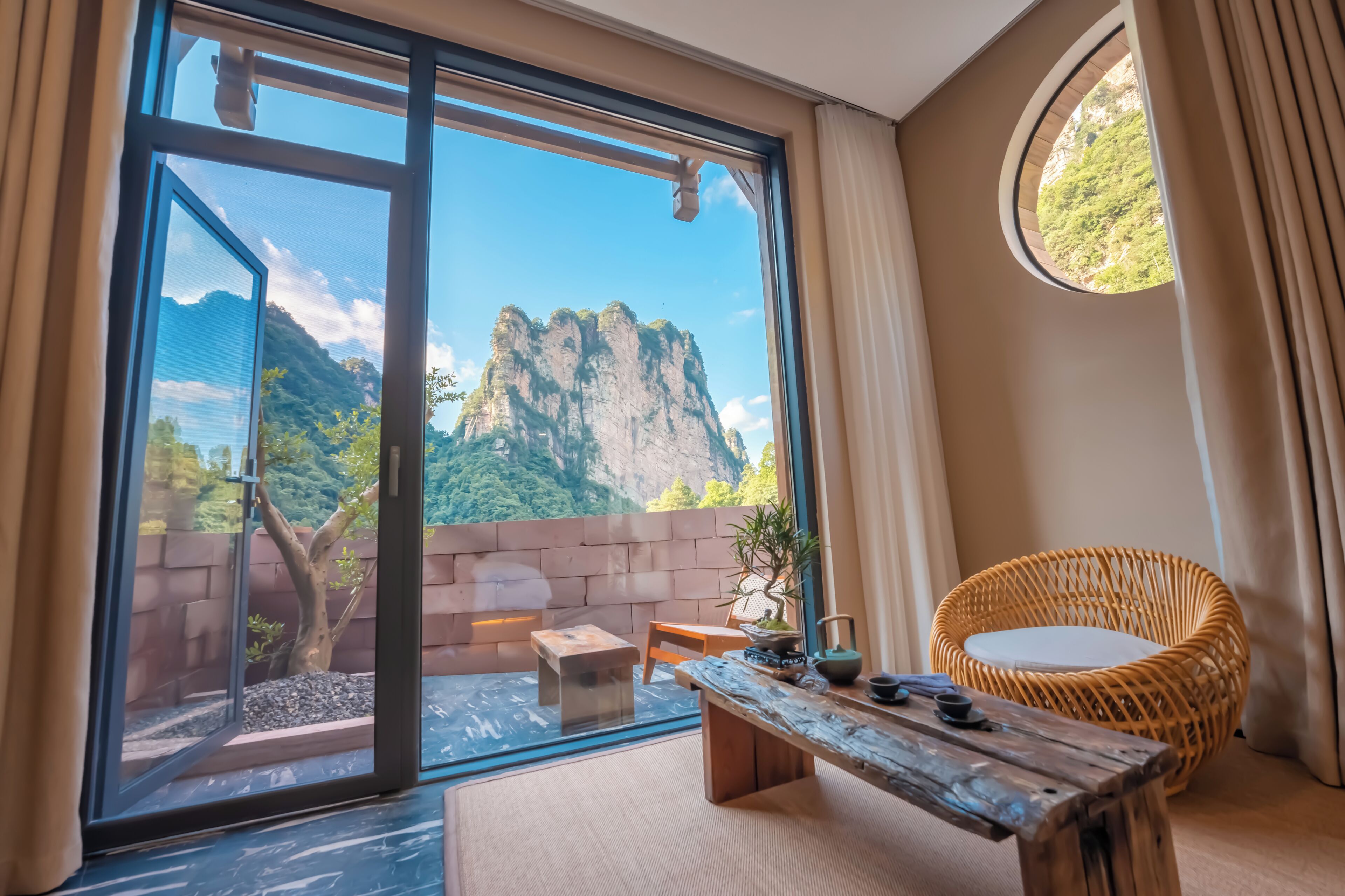 Una habitación con un ventanal grande, una silla de mimbre, una mesa de madera y vistas a las montañas.
