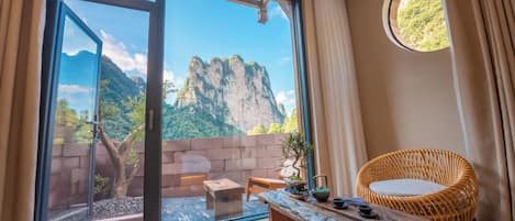 A room with a large window, a wicker chair, a wooden table, and a view of mountains.