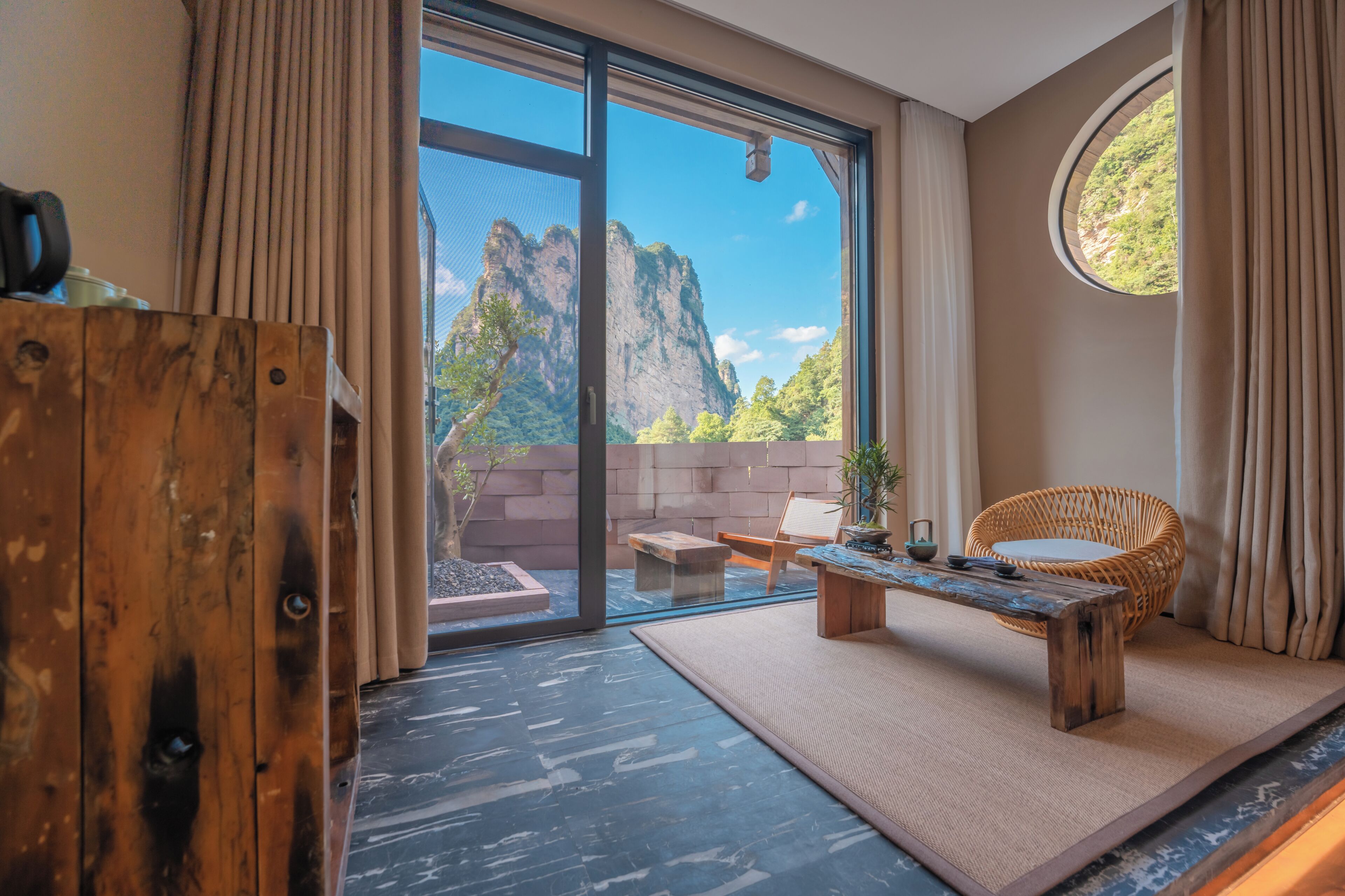 Habitación moderna con un amplio ventanal corredizo, una mesa de madera, una silla de mimbre y vistas a la montaña.