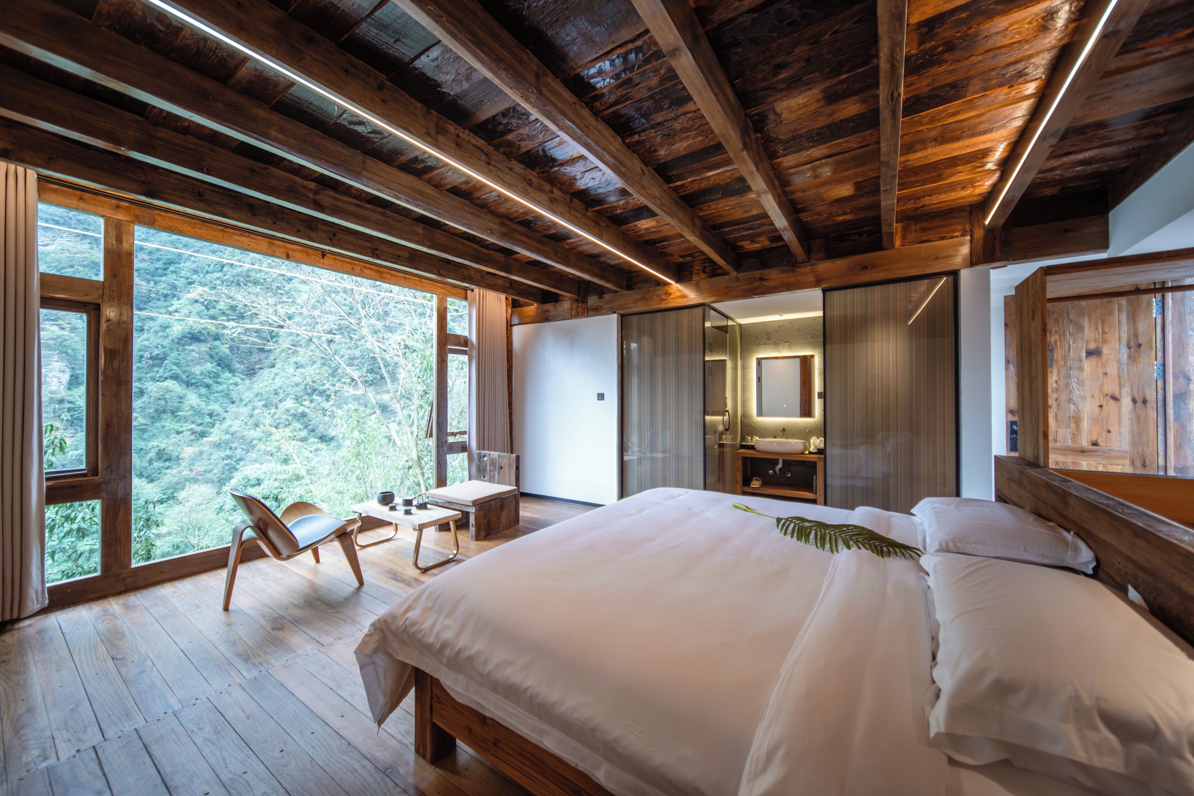 A bedroom with a large bed, wooden ceiling beams, a desk, and a view of the outdoors.