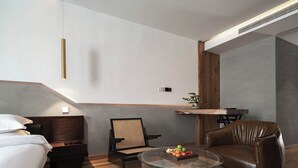 Quarto de hotel moderno com uma cama, um sofá de couro, uma mesa de centro redonda de vidro com frutas e uma pequena mesa lateral de madeira.