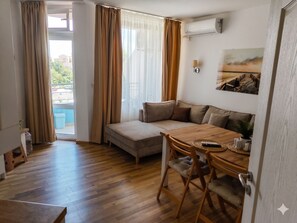Ein Zimmer mit Holzboden, einem beigefarbenen Sofa, einem Holzesstisch und Blick ins Freie durch ein Fenster mit Vorhängen.