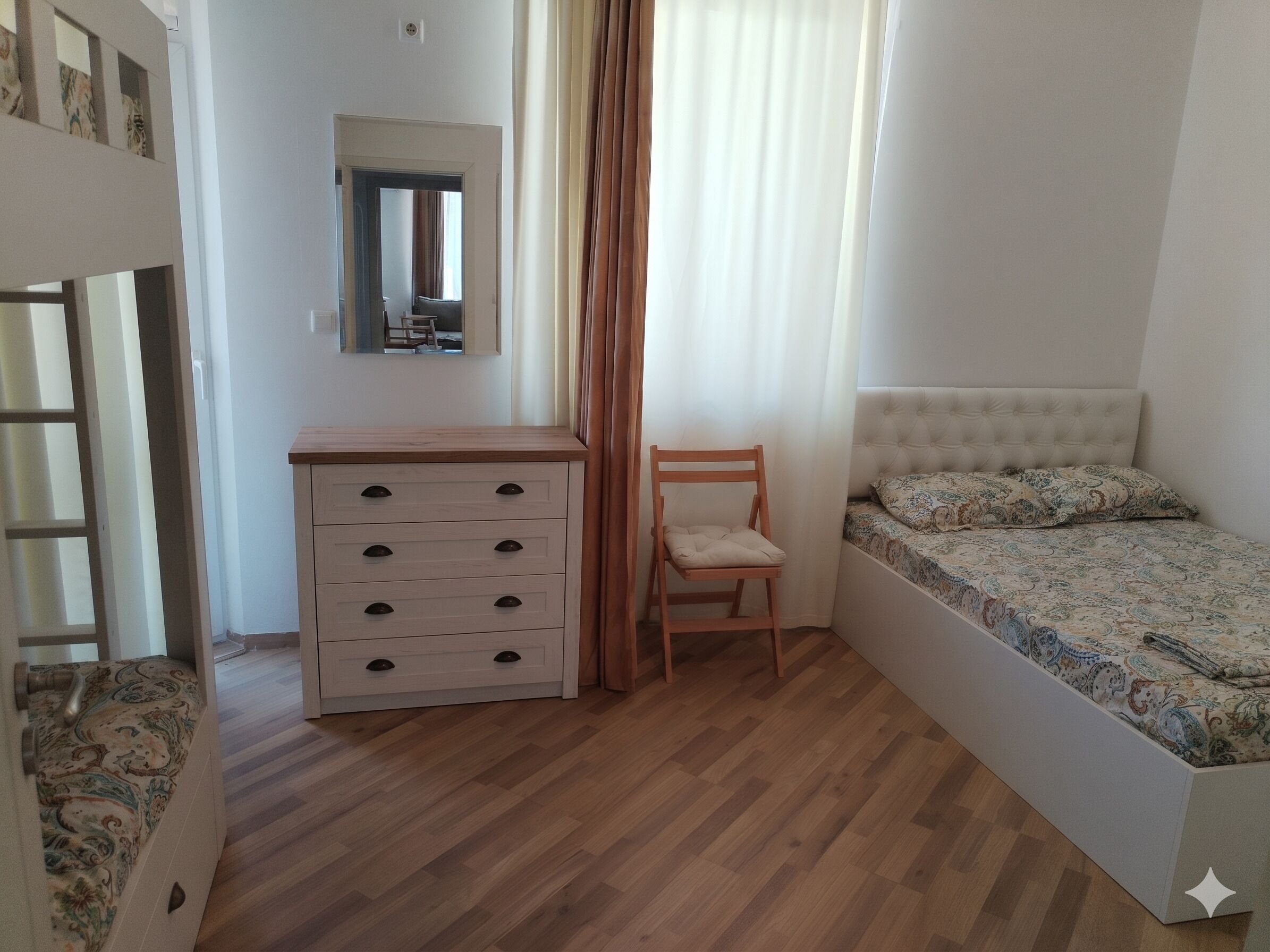 Ein Schlafzimmer mit einem hölzernen Schminktisch, einem Sessel, einem Bett mit gemusterten Betttextilien, einem Spiegel an der Wand und einem Fenster mit Vorhängen.