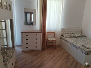 Ein Schlafzimmer mit einem hölzernen Schminktisch, einem Sessel, einem Bett mit gemusterten Betttextilien, einem Spiegel an der Wand und einem Fenster mit Vorhängen.
