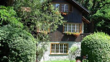 A two-story wooden house with a stone foundation, surrounded by dense greenery.