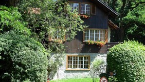 A two-story wooden house with large windows and a stone foundation, surrounded by lush greenery. - Rothmund, (Bad Ragaz), by Interhome (Bad Ragaz)