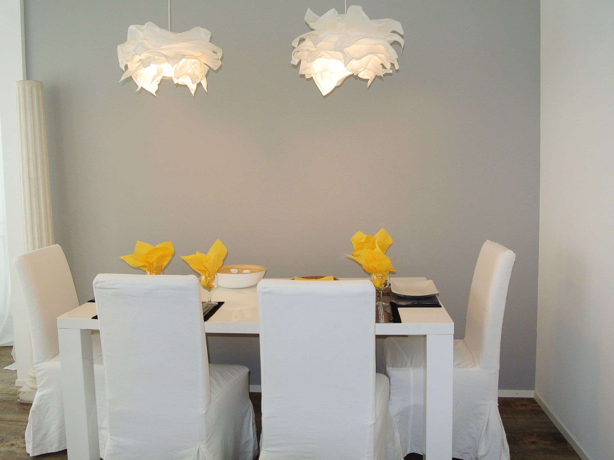 Una mesa de comedor con sillas blancas y servilletas amarillas, frente a una pared gris con dos lámparas colgantes blancas.