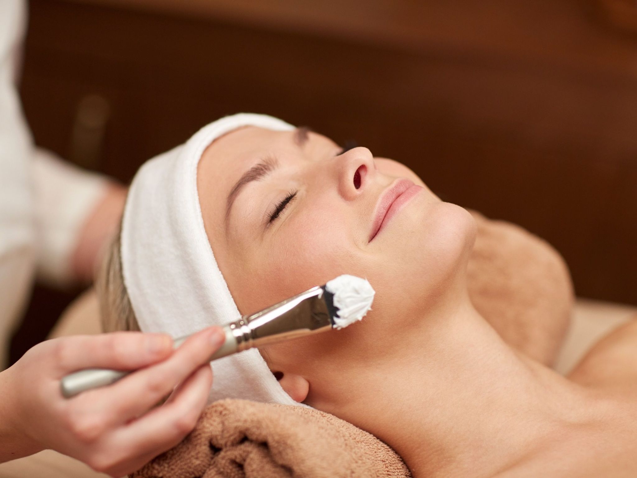 A person lying down with a towel, a brush applying a substance to their face, and a wooden background.