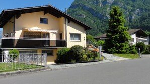 Un edificio de dos pisos con techo plano, balcón y montañas al fondo.