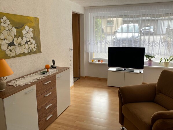 A living room with a sofa, a TV on a stand, a painting on the wall, and a window with blinds.