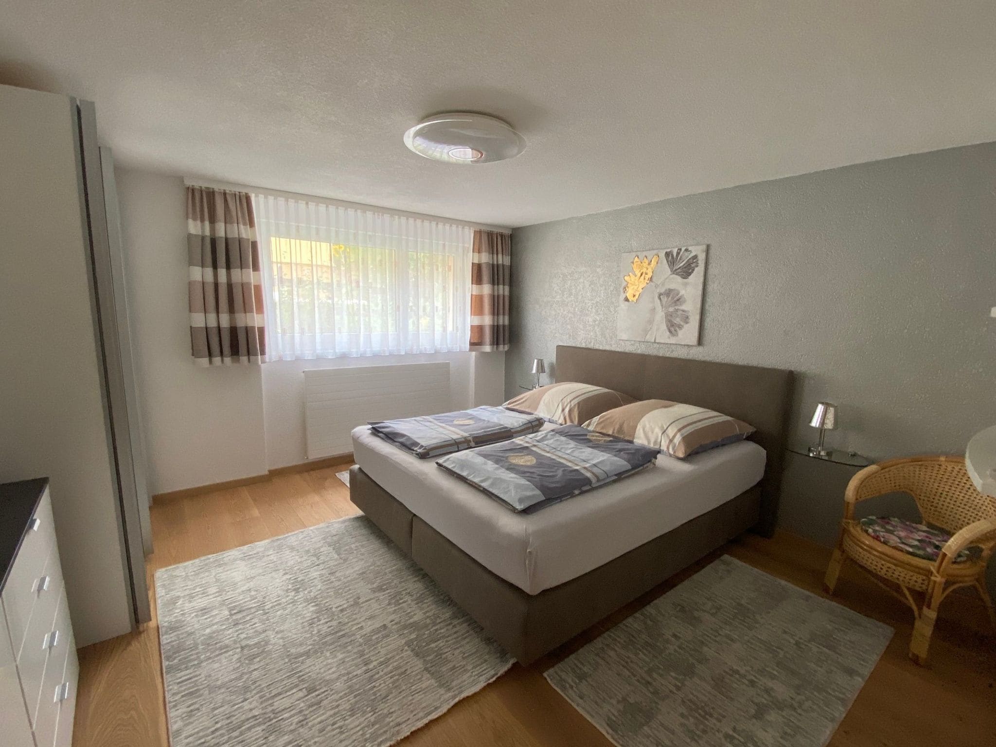 Ein Schlafzimmer mit einem Bett, zwei Nachttischen, einem Fenster mit Vorhängen, einem Wandbild und einem Sessel.