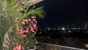 Ein Balkon mit einer Topfpflanze und Blumen, einem Geländer und Blick auf die Stadt bei Nacht.