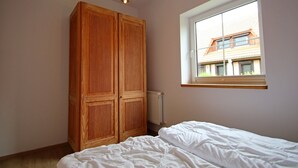 Un dormitorio con un armario de madera, una cama con ropa blanca, una ventana con vista a un edificio y una lámpara colgante.