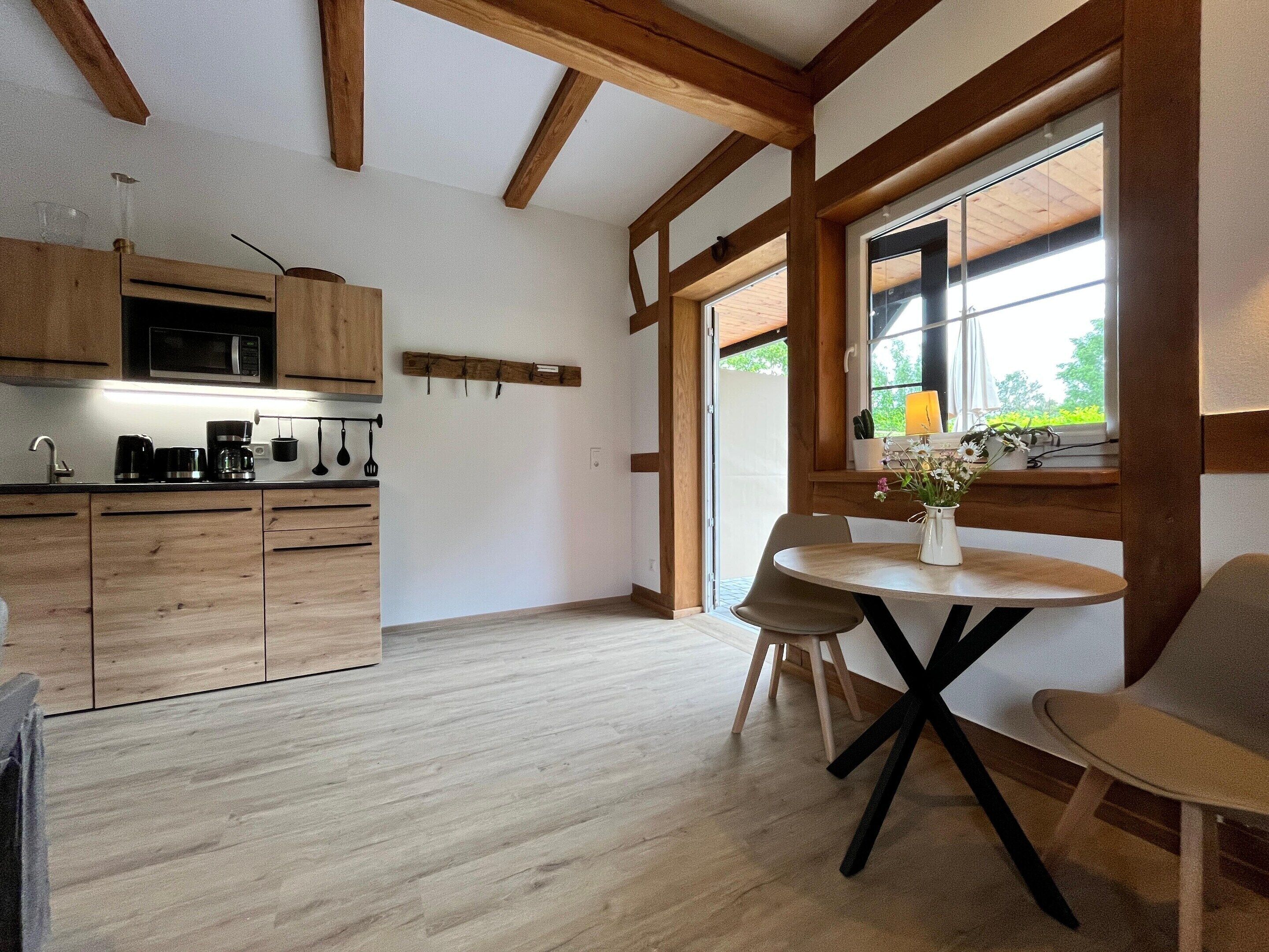 Eine moderne Küche mit Holzschränken, einer Mikrowelle und einer Kaffeemaschine. Ein Essbereich mit Tisch und Stühlen sowie ein Fenster mit Blick auf Grünflächen.