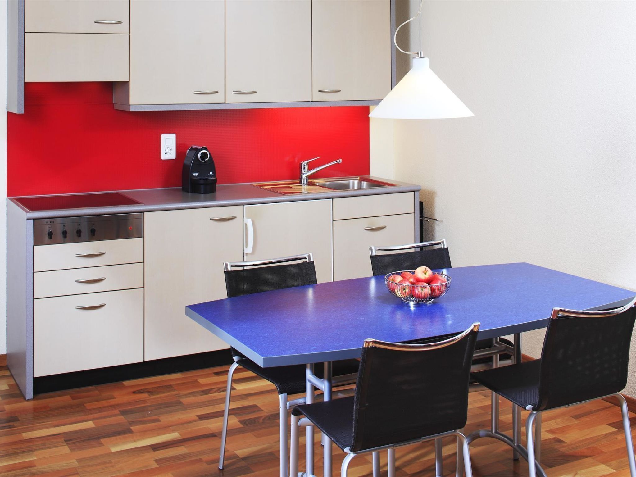Una cocina compacta con salpicadero rojo, un lavamanos, una cafetera y un comedor con una mesa azul y cuatro sillas.