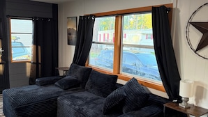 A room with a sectional sofa, a side table with a lamp, a window with curtains, and a view of the outside.