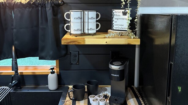A kitchen with a coffee maker, mugs, a kettle, and a sign on the wall.