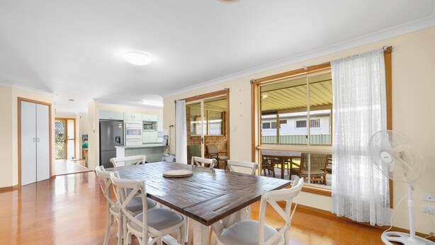 A dining area with a wooden table, white chairs, a fan, and a view of a building through a window. - Yannarie: 3  BR, 1.5  BA House in South West Rocks, Sleeps 7 (South West Rocks)