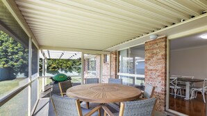 A covered patio with a round wooden table and wicker chairs, a brick wall, and a view of the backyard. - Yannarie: 3  BR, 1.5  BA House in South West Rocks, Sleeps 7 (South West Rocks)