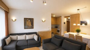 A modern living room with a sectional sofa, a smaller sofa, and a wooden coffee table. There is a kitchen area with wooden cabinets and a dining table with chairs. - Residenz Darianne Leukerbad # 5-bed apartment, Dusche und Bad, WC by Interhome (Leukerbad)