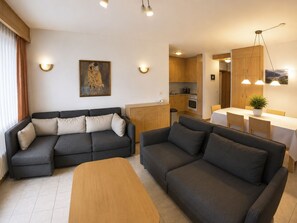 Ein modernes Wohnzimmer mit einer Eckcouch, einer kleinen Couch und einem Couchtisch aus Holz. Es gibt eine Küchenzeile mit Holdekor und einen Esstisch mit Stühlen.