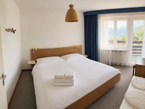 Ein Hotelzimmer mit einem großen Bett, einem hölzernen Kopfteil, einer Pendelleuchte, einem Fenster mit Vorhängen und Blick ins Freie.